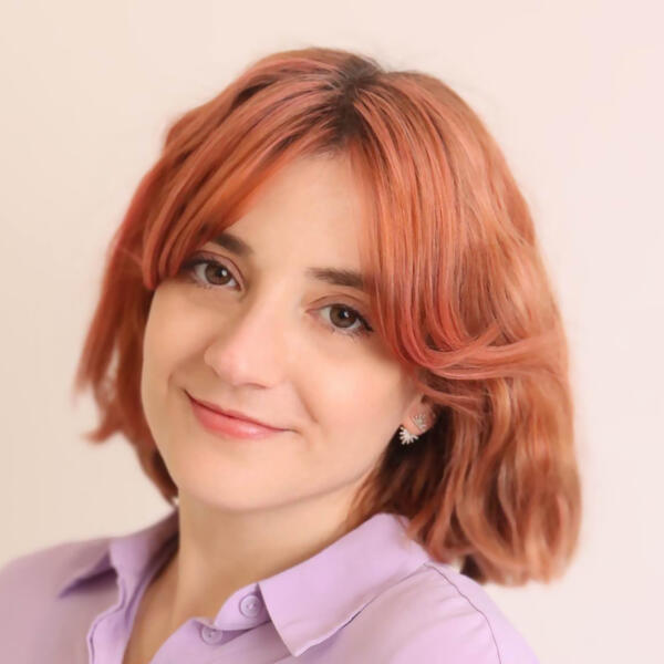 Headshot of Irene Silvera, with shoulder-length auburn hair, wearing a light lilac shirt, looking at the camera against a pale background